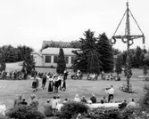 Folkdanslag har uppvisning vid midsommarstången på Kustsanatoriet Apelviken. En fiolspelare står för musiken. Patienter och personal åser uppvisningen på båda sidor om dansarna. Till vänster sitter en grupp rullstolsburna.