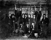 Jaktlag i en lada tillsammans med två upphängda, skjutna älgar. Männen håller i sina gevär och framtill ligger en jakthund. Troligen på Skällinge, som fotografen var ifrån