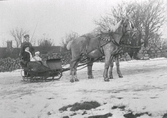Snöbild av ett hästekipage där kvinna och barn sitter under en fäll i släden och kusken står bakom dem. Hästarna har bjällror. Möjligen är bilden tagen på Kungsgatan i Varberg (se bildnr MR2_1037)