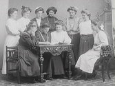 Tio unga kvinnor arrangerade i en scen där tre av dem sitter och läser vid ett bord (Se även bildnr MR2_1038)