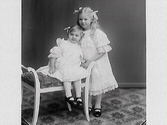 Barnporträtt av två flickor i spetsklänningar och rosetter i håret, systrarna Svea och Ingrid Hallberg.