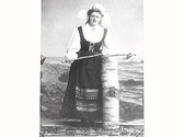 Kvinna i folkdräkt med en träpinne vid björkstubbe mot havsfond.