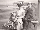 Familjebild, kvinna och man med flicka sittande mot fond med Varbergs fästning. (Se även bildnr MR2_1034)