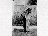 Liten pojke i kostym står leende i en trädgård och spelar fiol.