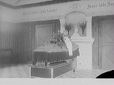 Fransprydd kista med blommor på katafalk vid enkelt altare, troligen i ett kapell eller frikyrka. Salen har omålade brädväggar, målad dekor upptill, rutigt golv och dörrar med kantställd rutdekor på ömse sidor om  (Glasplåten är skadad i högra nedre hörnet)