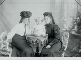 Porträtt av två kvinnor sitter med ett litet barn på ett bord mellan sig, beställt av Karstorp. Troligen är det tre generationer. Kvinnorna bär ovanlig mörka hattar med slöjor baktill.