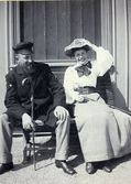 Fotografi från August Bondesons vänkrets. Man och kvinna på en bänk i solskenet intill en vägg. Han bär vegamössa och käpp, hon har resårskärp i midjan, stor halsrosett och håller i hatten.