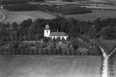 Flygfoto över Slöinge kyrka och kyrkogård med  omgivande odlingslandskap. På åkern nedtill står elstolpar.