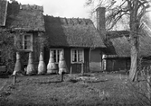 Ryggåsstuga fotograferad 1936, Larsagården Lustorp. Bilden visar fem sammanbyggda volymer av olika höjd, täckta med strå eller spån. Den lägsta delen är knuttimrad och har en hög skorsten. Framför huset står fem strutliknande bikupor av halm på en bänk.