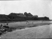 Vy från Kallbadhuset mot Varbergs fästning och Barnens badstrand i förgrunden. Till vänster ses ett några badhytter och barn leker på stranden.