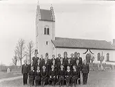 Mörkklädd konfirmandgrupp utanför Öxabäcks kyrka, Marks kommun. Prästen Hagman sitter i mitten.