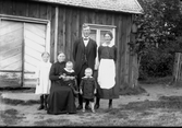 Familjebild med makar, tre barn och en äldre kvinna framför en byggnad med bred, tvåbladig port.