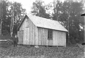 En man står vid en ny mindre byggnad med dörr och vindslucka på gaveln och ett stort fönster på långsidan. I förgrunden en åker, troligen med potatis.