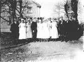 En samling män och kvinnor på en väg med en mangårdsbyggnad i bakgrunden. Kvinnorna är vitklädda med förkläden och hucklen. Kanske personal på en större gård eller en institution.