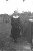 Flicka vid grind i konfirmationskläder med psalmboken i händerna. (Se även bildnr HB436, HB437, HB441, HB444)
