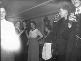 Ungdomar har kalas och står med dryckesglas i salongen. Vid vägguret står ett hembiträde. (Se även bildnr HB472)