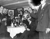 Ungdomar runt bordet på en fest utbringar en skål med skedar i glasen. På bordet står ett tänt ljus, en porslinskanna och flaskor. (Negativet skadat upptill. Se även bildnr HB466)