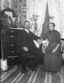 Ett par sitter vid ett bord och håller varandra i händerna. Fönstrets rullgardin är neddragen och på byrån intill står en spegel, så kallad pigtittare, en ljusstake och en flagga.