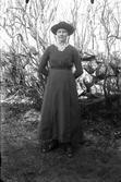 Ung kvinna vid en stengärdsgård. Hon bär hatt, hög spetskrage och sidoknäppt, lång klänning.