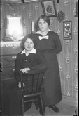 Två kvinnor i ett rum med jugendtapeter. Den ena kvinnan sitter på en pinnstol och den andra står bredvid. På bilden ses också en byrå med en spegel på, s k pigtittare.