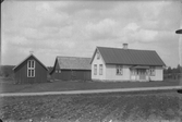 Lantgård med bostadshus och ekonomibyggnader. Den högra byggnaden har två stora fönster på gaveln. I förgrunden en plöjd åker längs den smala vägen.