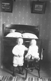 Bröderna Axel och Sten Andersson som små i Maås, Drängsered. De är fotograferade i en sängkammare med jugendtapeter. På sängens ena kudde ligger en hatt.