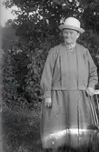 Kvinnoporträtt. Äldre kvinna i hatt står utomhus vid en stol.