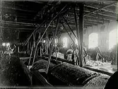 Fabriksinteriör där stora träkar dras runt med remdrift. Vid fönstren står  män och arbetar.