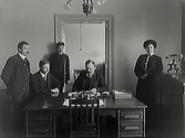 Kontor med tre män vid skrivbordet, en kvinna vid telefonen och en yngling som står vid dörrposten i bakgrunden. På bordet ligger olika föremål.
