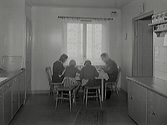 Vardagsbild där en familj med två barn sitter i köket och äter. Barnen sitter på pallar, föräldrarna på pinnstolar. Över diskbänken sitter hyllor med speceriburkar och en väggklocka är placerad ovanför dörröppningen till höger. Dörren till vänster leder sannolikt till ett skafferi.