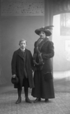 Ateljébild med kvinna och pojke stående i helfigur, båda iklädda ytterkläder. Kvinnan bär hatt och har händerna  istuckna i en muff. Pojken har en skärmmössa i höger hand. Beställare: Mia Andersson, Björketorp.