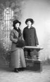 Ateljébild av två unga kvinnor stående i helfigur vid en rekvisitamur. Båda iklädda ytterkläder och pälshattar och dessutom har den ena kvinnan muff. Beställare Tekla Andersson.