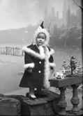 Flickporträtt av byggmästare Johannes Nilssons dotter i vinterkläder mot en fond med snöigt landskap. Kappan och mössan är pälsbrämade och bredvid henne på en rekvisitabalustrad står ett par leksaker; en katt och en fågel.