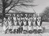 Klassfoto av flickor som gått en skolkökskurs. De är samlade utomhus i snön med en byggnad i bakgrunden.