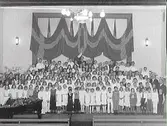 Stor barnkören framträder i aulan på Folkskolan/Rosenfredsskolan. På väggen bakom dem hänger draperade flaggor. (Se även bildnr GB2_3407)