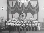 Gruppbild av en blåsorkester med gossar i Folkskolan/Rosenfredsskolans aula. Bakom dem hänger draperade flaggor. (Se även bildnr GB2_3406)
1:a raden från vänster: ?,?, Bertil Eliasson, ?, ?, Gunnar Svensson, Stig Hjalmarsson, Bertil Johansson, ?, ?, Erling Andersson, Inge Karlsson. 2:a raden: Lars Nilsson, ?, Bertil Nilsson, Sven Bertil Johansson, Thorvald Johansson, Gösta Nilsson, Ingvar Lenfors(?), Gösta Bengtsson, Karl Gunnar Strandberg. 3:e raden från vänster: Nils Appelgren, ?, ?, ?, Bertil Persson.