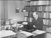 Museichef Albert Sandklef på kontoret i sin bostad i artillerikasernen på Varbergs fästning. Han sitter vid skrivmaskinen på skrivbordet, som är belamrat med böcker och dokument, med bokhyllor bakom sig.