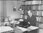 Museichef Albert Sandklef på kontoret i sin bostad i artillerikasernen på Varbergs fästning. Han sitter vid skrivmaskinen på skrivbordet, som är belamrat med böcker och dokument, med bokhyllor bakom sig.