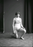 Ateljéfoto av kvinna i baddräkt och badmössa, stående på ett knä.