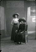 Ateljébild av två unga kvinnor i för tiden moderna bredbrättade hattar. Beställare: Ada Bengtsson.