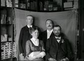 Tre män och en kvinna fotograferade i en butik, eller dess lager, troligen dess personal. En filt har hängts upp såsom bakgrund framför hyllorna där askar och lådor av olika slag förvaras. Beställare: John Högberg.