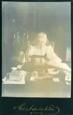 Porträtt av Gustafva Adelsköld (f. Brolin) (1844-1940) vid en symaskin och ett bord med böcker, sytillbehör mm. Fotograferad av hennes man Claes Adelsköld.