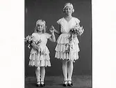 Syskonbild av två flickor i sommarskrud med blomdiadem, volangkjolar och blombuketter. Kamrer Bramstång beställde bilderna, sannolikt barnens far.