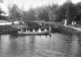 Sju personer i en roddbåt vid kanalen, Gröndal, Norrlandet, Gävle kommun 1902. Tre personer står på stranden.