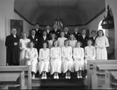 Konfirmandgrupp i Sibbarps kyrka med prästen Starke till vänster. I en nisch till höger står dopfunten med en ovanlig gammal krona hängande ovanför, skuren i trä, bemålad och med inskription.