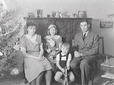 Makar med två barn i hemmet vid julgranen. Dottern håller i en docka och pojken sitter på en gunghäst.