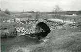 Gammal landsvägsbro med ett valv och smidda järnräcken nära Älvsåkers kyrka. I bakgrunden ses gårdsbebyggelse.