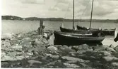 Sandö hamn med ett par förtöjda ekor och segelbåtar vid en liten stenpir. Där sitter en kvinna.