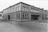 Exteriör av byggnaden i hörnet Torggatan-Östra långgatan. Vid Torggatan till höger syns affären Arons mattor och utmed Östra Långgatan till vänster syns Iris bageri. Bilden tagen mot sydväst under 1980-talet.