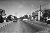 Gata genom Vallberga med bilar parkerade på vänster sida, eftersom högertrafiken ännu inte hade införts. Till vänster ses ett bageri och här finns även glassförsäljning och en bensinstation. Längs vägen står el- och telefonstolpar.
